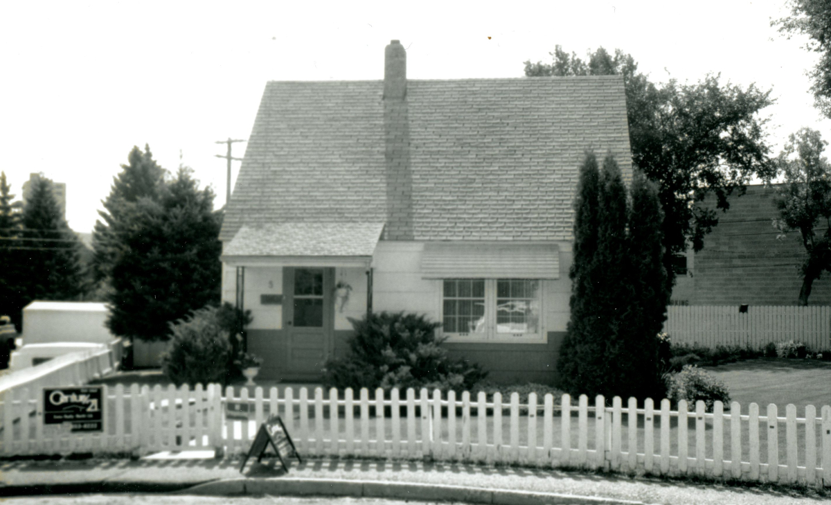 A single family house with picket fence in front, brick wall of another building behind the backyard, and flour mill in the distance.